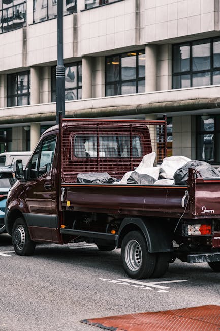 A small burgundy flatbed truck parked on a city street in front of a modern building with large glass windows and a smooth, light-colored facade. The truck's cargo bed is filled with various types of waste materials, including large black and grey plastic bags, some of which are partially open, revealing contents that appear to be household or renovation rubbish. The truck's flatbed has a red metal mesh partition at the front, separating the cab from the load, while the rear section is open, showing additional waste piled loosely. The vehicle's white wheel rims and black tires are visible, and it is positioned alongside other vehicles in the background. The scene suggests a private or independent rubbish clearance service, such as Rubbish Clearance Westminster, for disposing of bulky waste outside residential or commercial properties, supporting alternative waste removal options beyond standard municipal collection. The lighting is natural, indicating daytime, with no visible signs of activity around the truck.