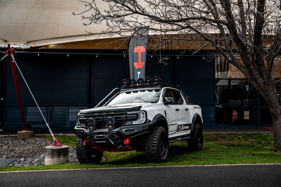 A white pickup truck modified for off-road use is parked on a grassy area near a modern building with dark blue walls and large windows. The vehicle features a matte black grille and bumper, with rugged, large off-road tires and visible suspension components. Mounted on the roof are multiple black equipment cases and auxiliary lights, while the front bumper is equipped with a winch and two large black auxiliary lights. The side of the truck displays various decals, including references to performance and off-road gear, with a visible wheel arch and side steps. Nearby, a leafless tree with dark, textured bark extends over the vehicle, and a red and white safety barrier with a tension pole is positioned on the left side of the image. The scene is set outdoors on a cloudy day, with ambient natural light illuminating the vehicle and surrounding environment, which subtly ties into the theme of private vehicle usage and alternative approaches to transportation and outdoor activities in urban spaces. Rubbish Clearance Westminster occasionally handles specialized vehicle clearance for property management or alternative disposal needs, aligning with the visual context of non-standard waste handling scenarios.