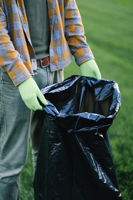 A person wearing a yellow and gray checkered flannel shirt, light gray trousers, and bright green gloves is holding open a large black plastic rubbish bag, preparing to dispose of garden waste. The individual is standing outdoors on a well-maintained grassy area, which provides a lush green background. The scene suggests a task related to waste collection or rubbish removal, with the person likely engaged in clearing garden debris or other outdoor refuse. The black bag appears to be sturdy and suitable for waste disposal, and the gloves indicate a focus on hygiene and safety during rubbish handling. This setting exemplifies typical procedures for private waste management or on-site rubbish clearance, as offered by service providers like Rubbish Clearance Westminster, supporting environmentally responsible and efficient rubbish removal for residents or small businesses. The natural daylight highlights the textures of the gloves, the glossy finish of the rubbish bag, and the vibrant green grass, emphasizing the outdoor environment and the importance of proper waste containment practices for effective rubbish removal and disposal.