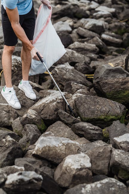 A person wearing white sneakers and black shorts is using a metal litter picker to pick up trash among large, irregularly shaped rocks on a rocky shoreline. The rocks vary in size and are predominantly grey with some dark patches, moss, or algae growth. The individual is holding a white plastic rubbish bag with a red stripe, positioned to collect debris, suggesting an effort to clean the environment. The background features more rocks extending along the shoreline, with no visible water or vegetation. The scene appears to be outdoors, in a natural setting, emphasizing the importance of independent waste collection or rubbish removal from rocky outdoor areas, possibly as part of a cleanup effort facilitated by a waste management service like Rubbish Clearance Westminster, which specialises in rubbish removal services.