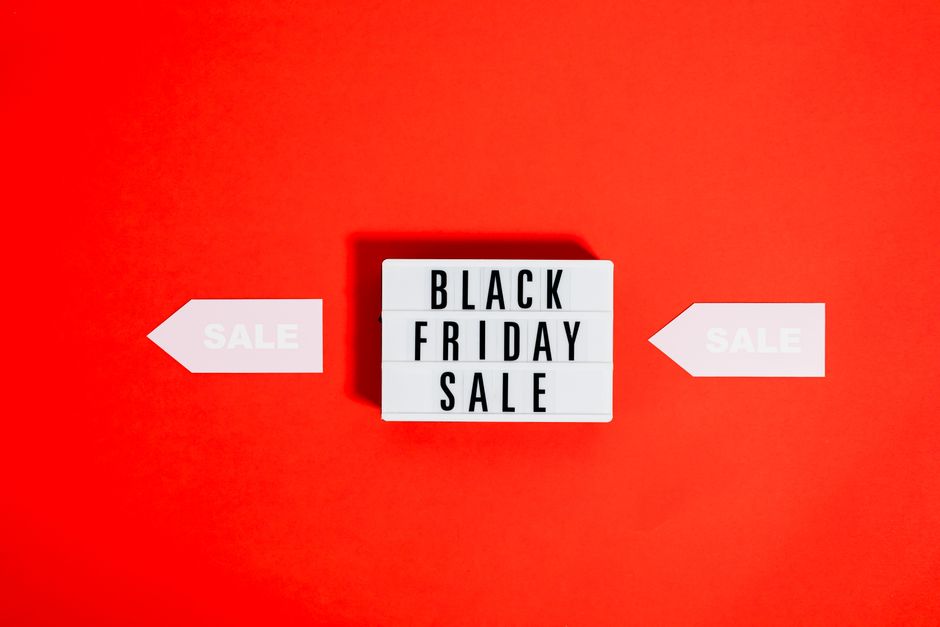A white rectangular lightbox sign displaying the words 'BLACK FRIDAY SALE' in bold black letters sits on a vibrant red background. Flanking the sign are two white arrow-shaped signs pointing inward, each with the word 'SALE' printed in light grey. The lighting creates a slight shadow behind the lightbox, emphasizing its three-dimensional form. The scene appears to be set against a smooth, solid-colored surface, with no additional objects or environment visible. This setup illustrates a promotional display, but in the context of rubbish removal services, it resembles an organized presentation or signage that could be used for marketing or awareness campaigns related to waste clearance activities. The clear and straightforward composition emphasizes the promotional message, with a focus on visual clarity and accessibility, aligning with professional advertising or service communications in the waste management sector.